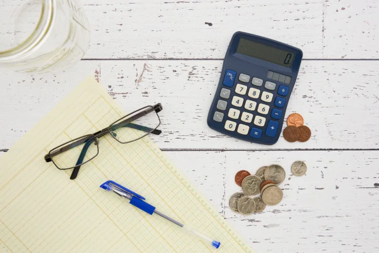 Calculator, reading glasses, coins and a ledger pad on a white wooden table