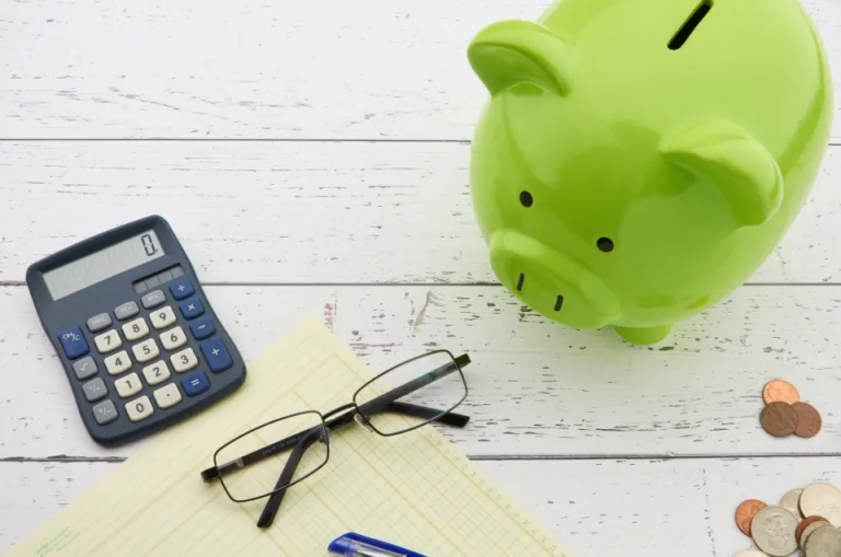A green piggy bank, a calculator, reading glasses and a few coins on a white table