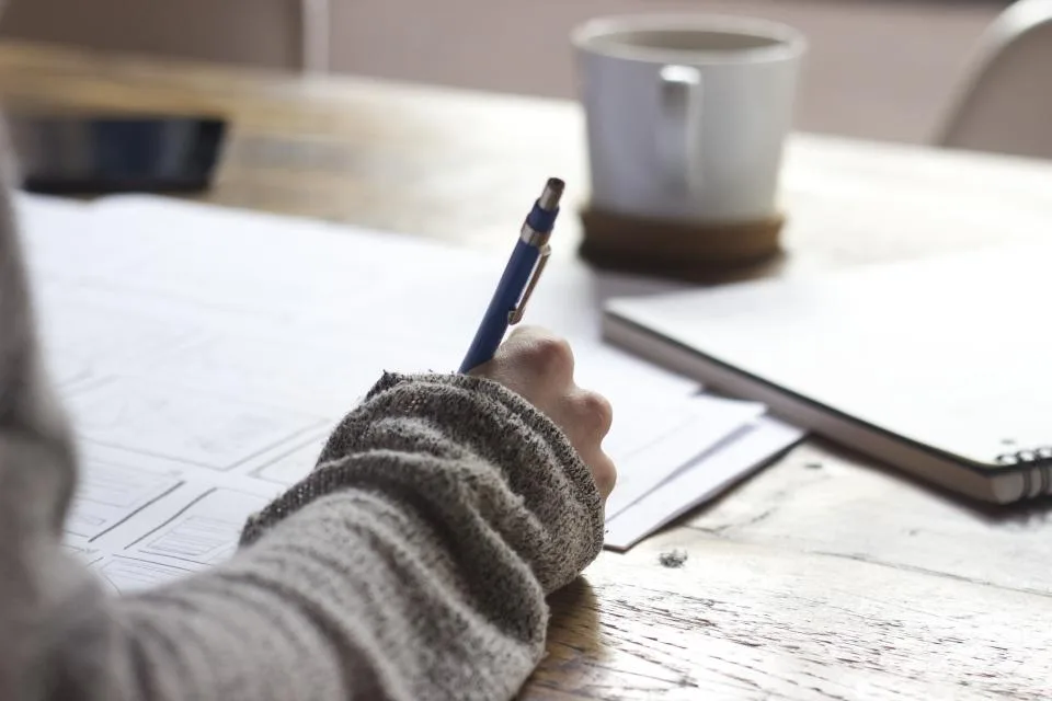 A hand writing notes with a pen at a wooden table beside a coffee mug