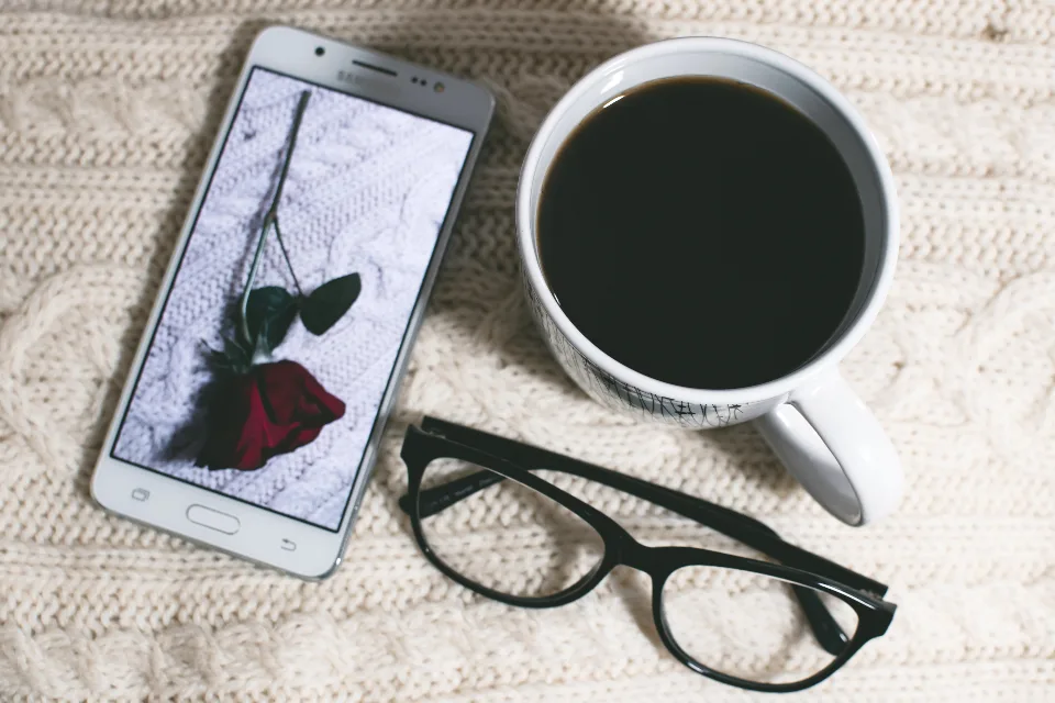 A smartphone, a cup of black coffee and reading glasses on a cream knit blanket