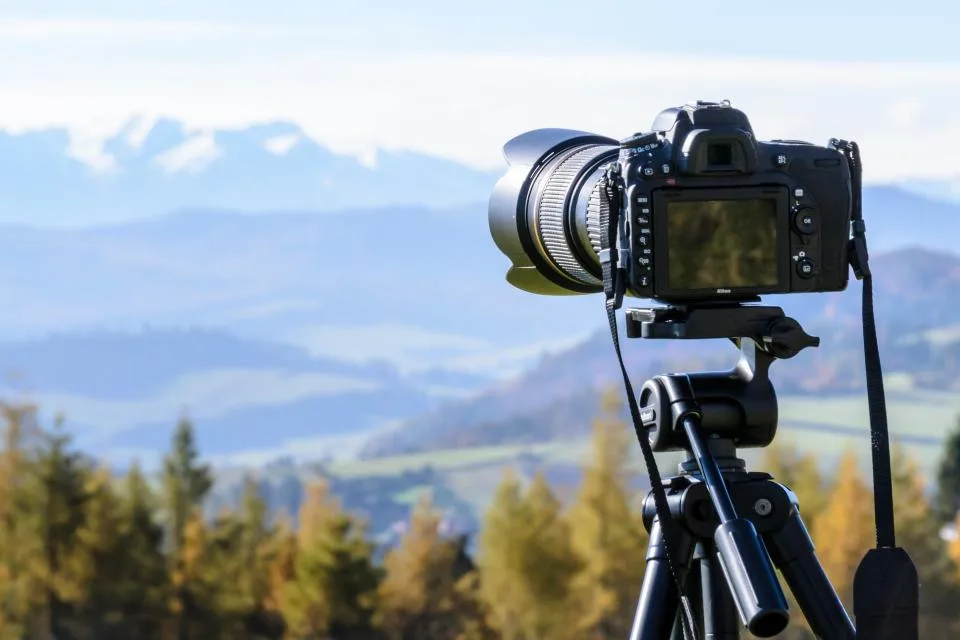 A camera on a tripod pointed at a valley of autumn trees and distant mountains