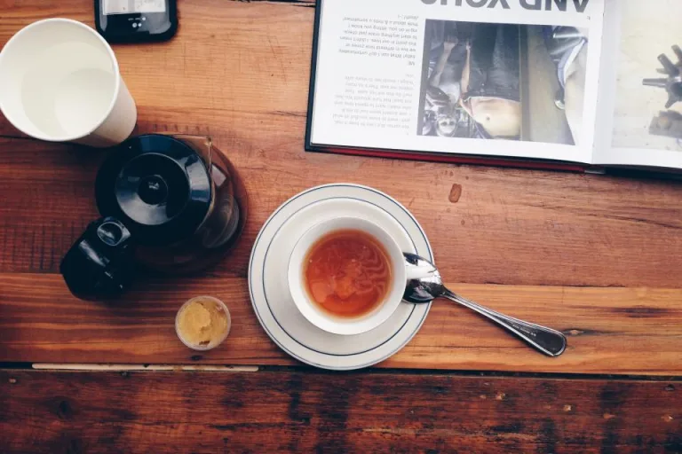 A cup of tea and a pot on a wooden table beside an open magazine