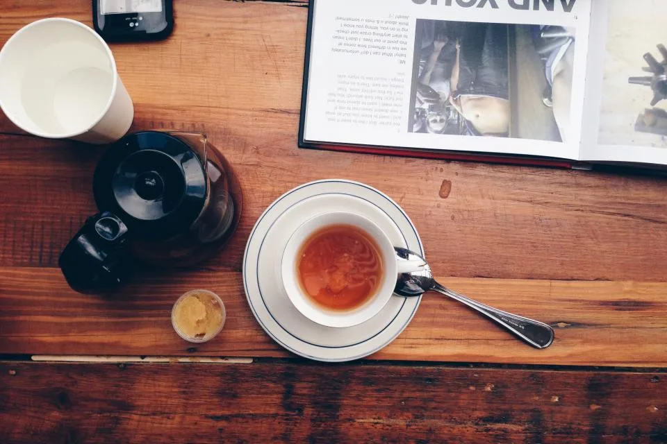 A cup of tea and a pot on a wooden table beside an open magazine