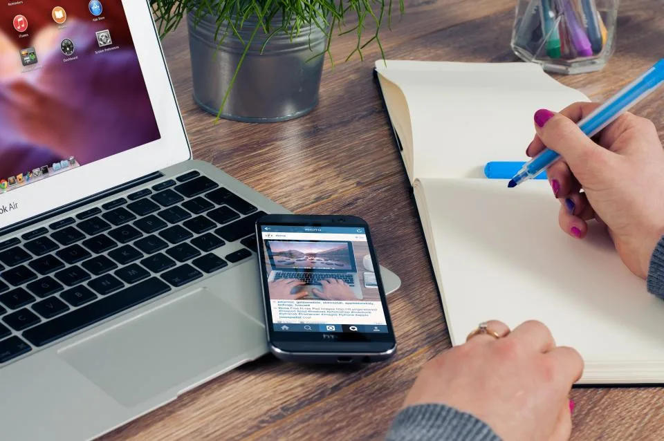 A laptop, phone and open notebook on a wooden table with a hand writing notes