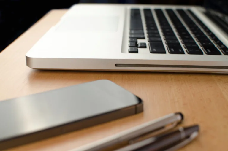 A laptop, a smartphone and two pens on a wooden desk