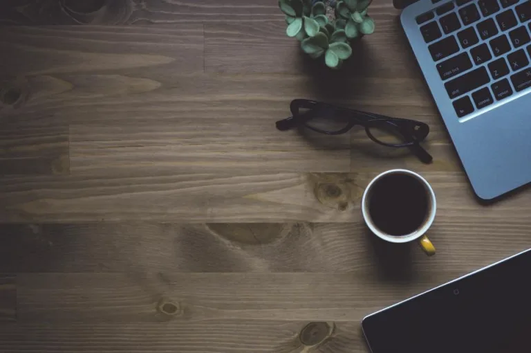 A laptop, a cup of coffee and reading glasses on a wooden table
