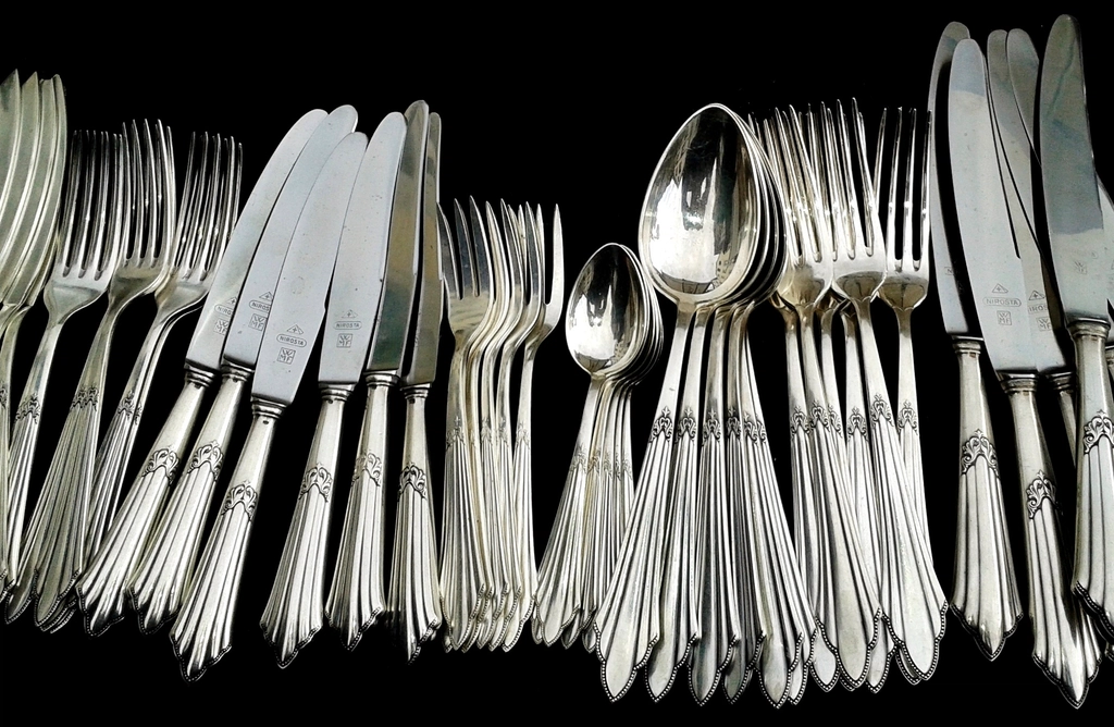 Rows of old silver forks, knives and spoons laid out on a dark background