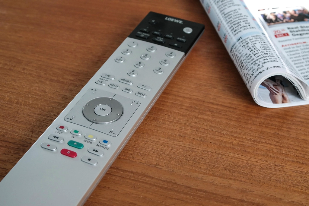A TV remote control lying on a wooden table next to a folded newspaper