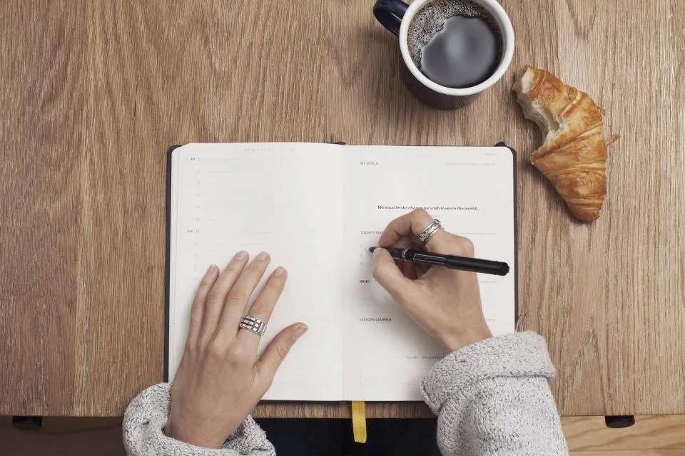 Hands writing in an open notebook on a wooden table beside a cup of coffee