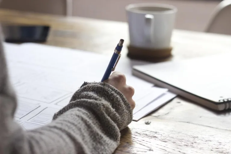 A hand writing notes with a pen at a wooden table beside a coffee mug